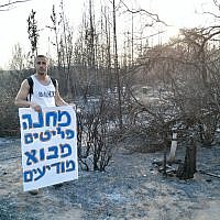 A resident stands next to burned grounds in Mevo Modi'im holds a sing that reads "Mevo Modiim Refugee Camp" in protest of the government's lack of support for the moshav, May 30, 2019. (courtesy Inbal Mustacchi)