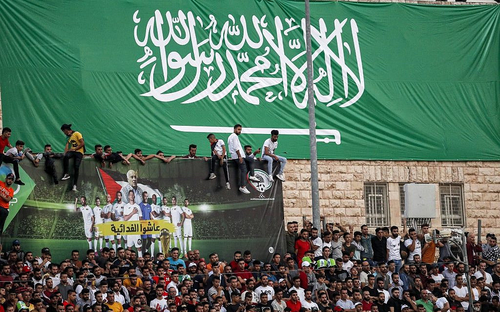 Football fans stand beneath a giant banner depicting the Saudi national flag as they attend the World Cup 2022 Asian qualifying match between Palestine and Saudi Arabia in the town of al-Ram in the West Bank on October 15, 2019. (Ahmad GHARABLI/AFP)