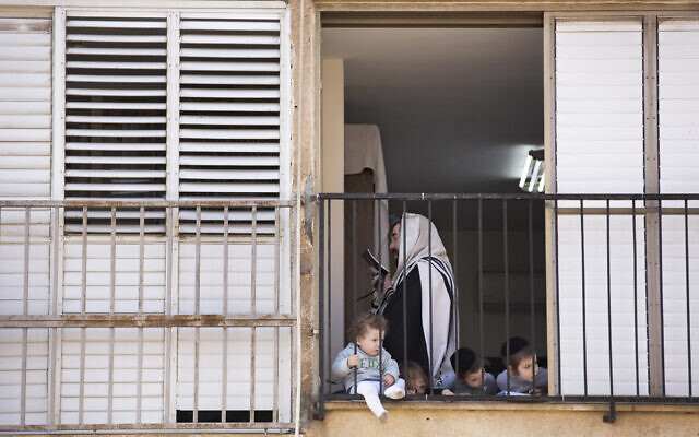 An ultra-Orthodox man prays the morning prayer in his apartment as his children look through the window during a lockdown imposed to slow the spread of the coronavirus, in Bnei Brak, April 8, 2020 (AP Photo/Oded Balilty)
