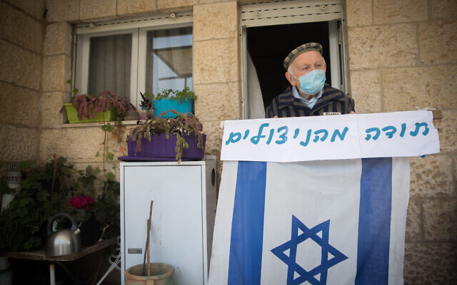Illustrative: Joseph Kleinman, a 90-year-old Holocaust survivor who survived Auschwitz and Dachau Nazi death camp wearing a face mask and holding an Israeli flag at his porch in Jerusalem, during the Holocaust Remembrance Day, April 21, 2020. (Yonatan Sindel/Flash90)