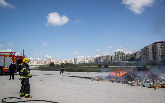 Israeli firefighters at a ceremony burning chametz, or leaven products, collected by the Jerusalem municipality in a final preparation before the Passover holiday, near the disused runway at the abandoned Atarot Airport, north of Jerusalem, on April 8, 2020. In the background are high-rises in the Kafr Aqab Palestinian neighborhood -- an area formally part of municipal Jerusalem but beyond the security barrier. (Yonatan Sindel/Flash90)
