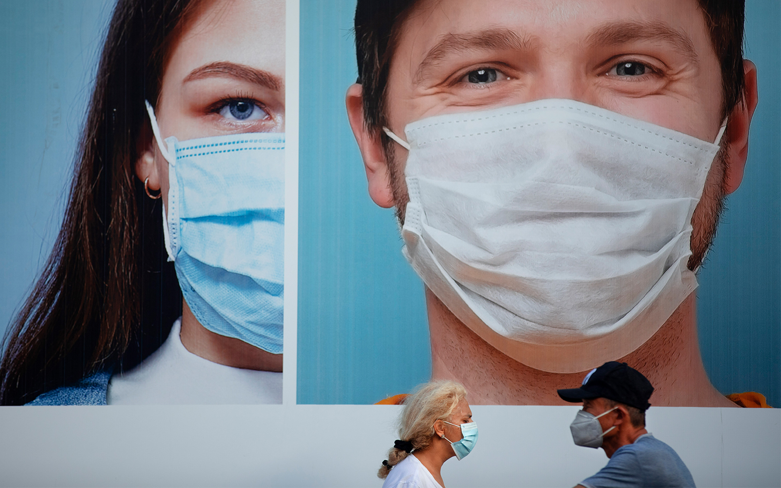 People wearing protective face masks pass a billboard in Ramat Gan, near Tel Aviv, July 16, 2020. (AP Photo/Oded Balilty)