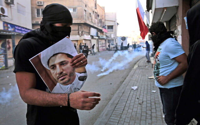 In this Jan. 3, 2015 file photo, a masked Bahraini anti-government protester holds a picture of jailed Shiite cleric Sheik Ali Salman, the head of the opposition al-Wefaq political association, as riot police fire tear gas canisters during clashes in Bilad Al Qadeem, Bahrain. (AP Photo/Hasan Jamali, File)