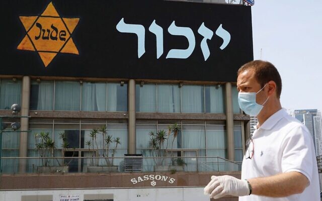 Illustrative: A man in a mask and protective gear stands near a banner depicting a Holocaust-era yellow Star of David, in Tel Aviv on April 21, 2020. (Jack Guez/AFP via Getty Images)