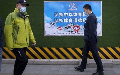 People wearing face masks walk past a poster promoting the 2022 Winter Olympics that reads 'Carry forward the Chinese sports spirit, promote sportsmanship,' in Beijing, on December 8, 2021. (AP Photo/Mark Schiefelbein)