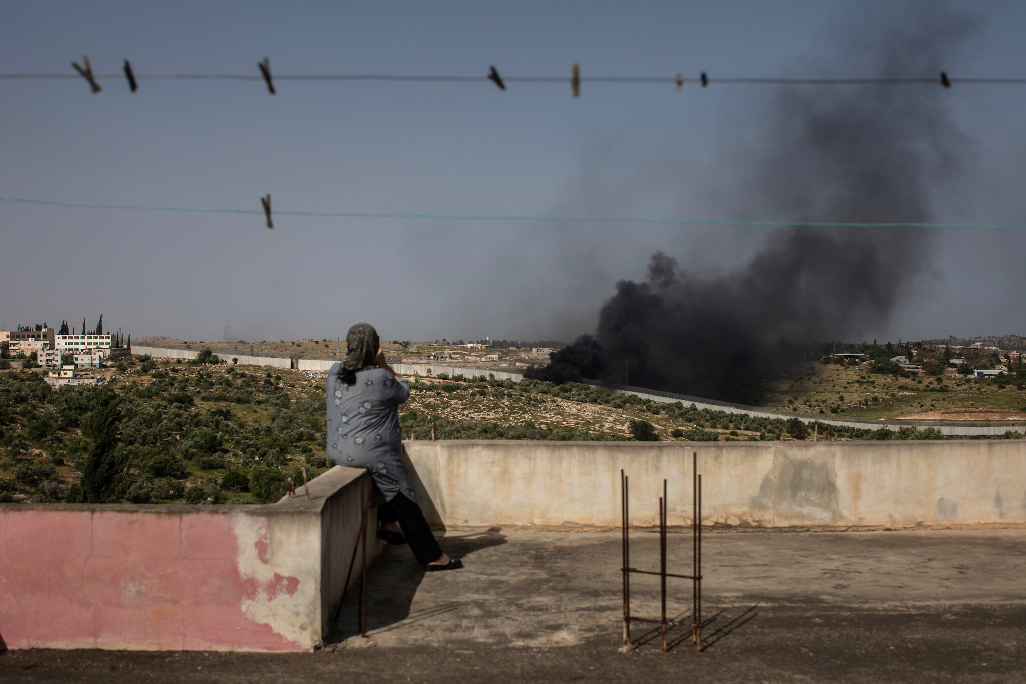Maysoon Sweity, a 54-year-old Palestinian from Beit Awwa, sits on the roof of her home and watches as e-waste and garbage burns near the concrete wall separating her village from Israel on April 26, 2019. (Tamir Kalifa)
