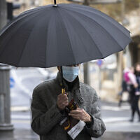 A man wearing a mask takes cover from the rain on Jaffa Road  in the city center of Jerusalem on December 21, 2021. (Olivier Fitoussi/Flash90)