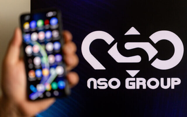 Illustrative. A man holding his phone with NSO Group logo on a computer screen at the background, in Jerusalem, on February 7, 2022. (Yonatan Sindel/Flash90)