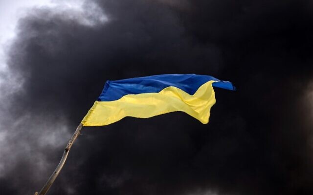 A Ukrainian national flag waves while smoke rises after Russian attacks hit a fuel storage facility in the city of Kalynivka, on March 25, 2022. (Photo by FADEL SENNA / AFP)