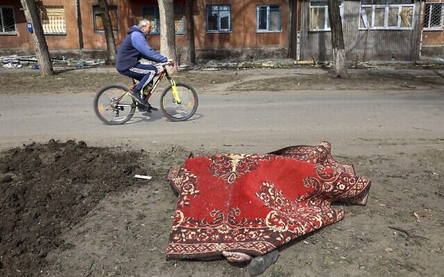A man rides his bicycle past a body covered by a rug after fighting on the outskirts of Mariupol, Ukraine, in territory under control of the separatist government of the Donetsk People's Republic, on March 29, 2022. (AP Photo/Alexei Alexandrov)