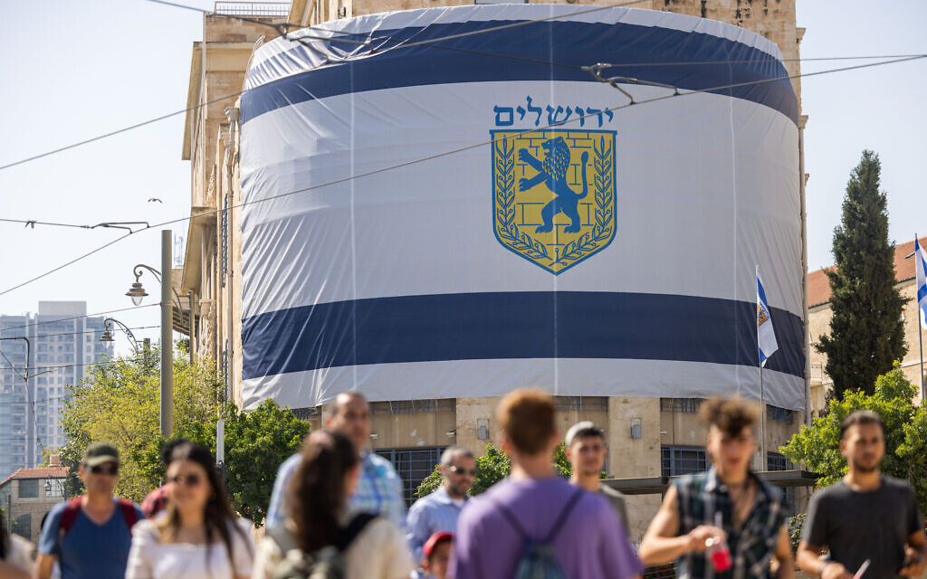 People walk past a giant Jerusalem Municipality flag hung across from the Old City on May 26, 2022, ahead of Jerusalem Day. (Yonatan Sindel/Flash90)