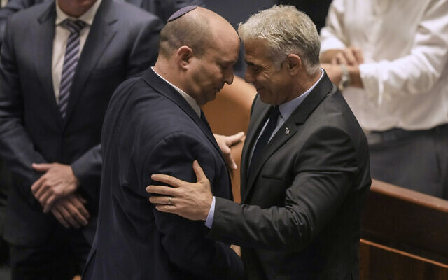 Outgoing Prime Minister Naftali Bennett, left, and incoming prime minister Yair Lapid embrace in the Knesset after the passage of a bill to dissolve the parliament and hold new elections, June 30, 2022 (AP Photo/Ariel Schalit)