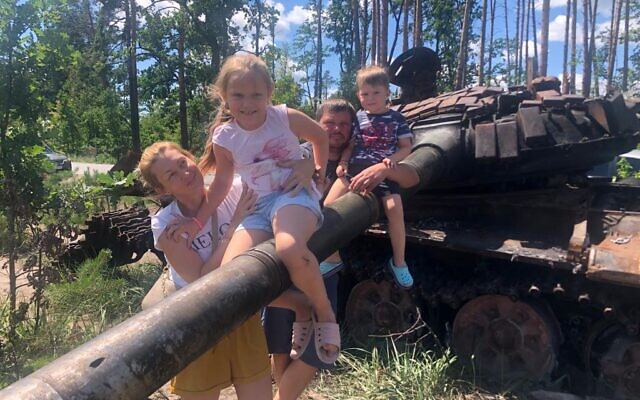 Olesei (L) and Ruslan poses with their children in front of a destroyed Russian tank outside of Kyiv, July 22, 2022 (Lazar Berman/The Times of Israel)