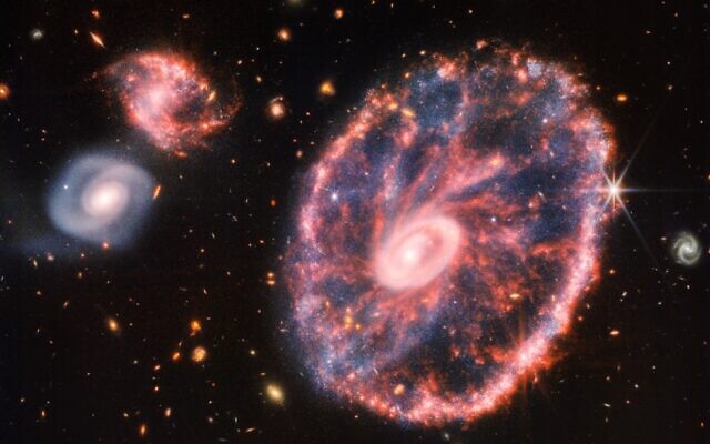 A large pink, speckled galaxy resembling a wheel with a small, inner oval, with dusty blue in between on the right, with two smaller spiral galaxies about the same size to the left against a black background. (NASA, ESA, CSA, STScI)