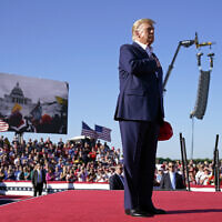 As footage from the January 6, 2021, insurrection at the US Capitol is displayed in the background, former US president Donald Trump stands while a song, "Justice for All," is played during a campaign rally at Waco Regional Airport, March 25, 2023, in Waco, Texas. (AP Photo/Evan Vucci)