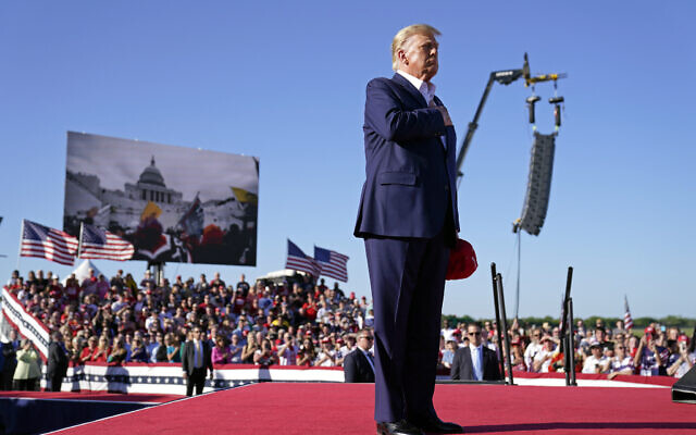 As footage from the January 6, 2021, insurrection at the US Capitol is displayed in the background, former US president Donald Trump stands while a song, "Justice for All," is played during a campaign rally at Waco Regional Airport, March 25, 2023, in Waco, Texas. (AP Photo/Evan Vucci)