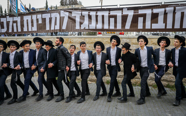 Haredi men and teens dance in front of a sign reading 'We must resist against a halachic state' as protesters rally outside the home Shas leader Aryeh Deri in Jerusalem, March 23, 2023 (Flash90)