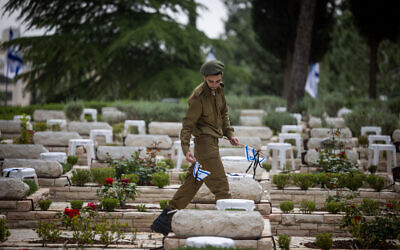 An Israeli soldier places flags, flowers and candles on graves of fallen soldiers at the Mount Herzl Military Cemetery in Jerusalem, April 23, 2023, ahead of Memorial Day. (Yonatan Sindel/Flash90)
