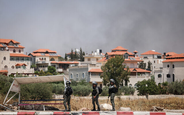 Smoke rises behind IDF soldiers at the entrance to Turmus Ayya, as dozens of settlers reportedly rampage through the West Bank village, June 21, 2023 (Yonatan Sindel/Flash90)