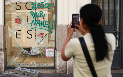 A pedestrian uses a smartphone to take footage of a damaged store facade with a message reading 'Justice for Nahel' in Marseille, southern France on July 1, 2023, after a fourth consecutive night of rioting in France over the killing of a teenager by police. (Clement Mahoudeau/AFP)