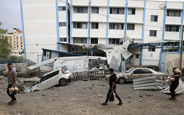 Illustrative: Palestinian men carrying bread walk past damaged cars and debris in front of a school run by the United Nations Relief and Works Agency for Palestine refugees (UNRWA) following Israeli airstrikes in Gaza City on October 9, 2023. (Mahmud HAMS/AFP)