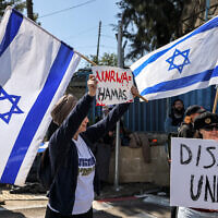 A protester holds an Israeli flag and a sign while standing with others gathering outside the West Bank field office of the United Nations Relief and Works Agency for Palestine Refugees (UNRWA) in Jerusalem on March 20, 2024. (AHMAD GHARABLI / AFP)