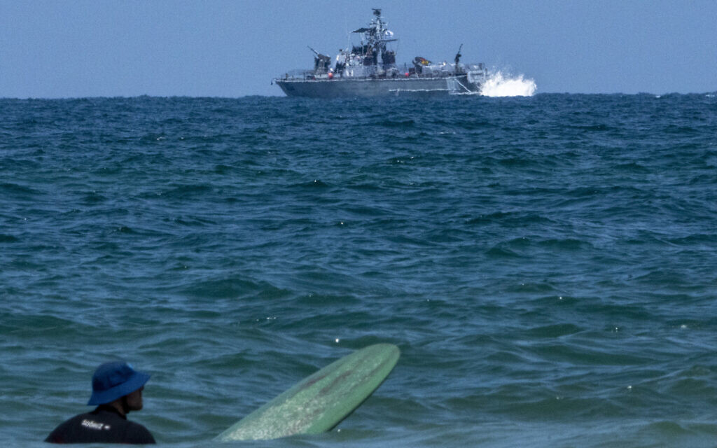 A surfer waits for wave while an Israeli military naval ship patrols the Mediterranean sea off the coast of Hadera, April 14, 2024 (AP Photo/Ariel Schalit)