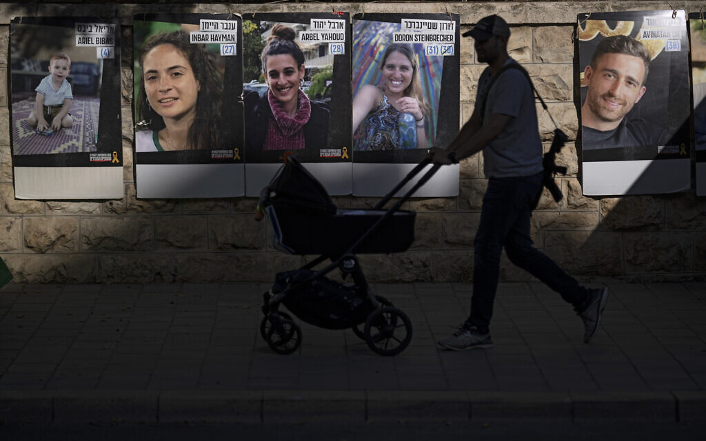 Carrying an weapon, a man pushes a baby stroller past a wall with photographs of hostages who were kidnapped during the Oct. 7 Hamas cross-border attack in Israel, in Jerusalem, Sunday, June 9, 2024. (AP/Leo Correa)